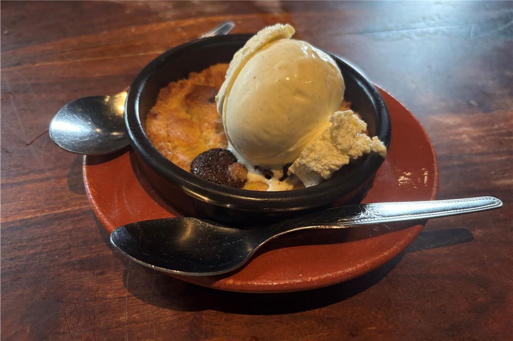 „O Mira“ im Restaurant-Check: Was kann der „Carlos“-Nachfolger? Auf dem Foto ist ein Dessert zu sehen, bestehend aus einem gebackenen Cookie in einer kleinen schwarzen Schüssel, garniert mit einer Kugel Vanilleeis und einem Schokoladenstück. Das Dessert wird auf einem Teller serviert und von zwei Löffeln begleitet.