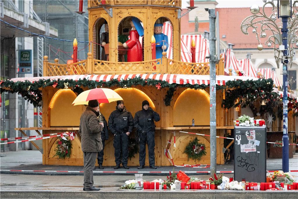Anschlag auf Magdeburger Weihnachtsmarkt: Fünf Tote, 200 Verletzte - alle Infos im Liveblog Ein Passant mit Regenschirm und Polizisten stehen vor dem Weihnachtsmarkt.