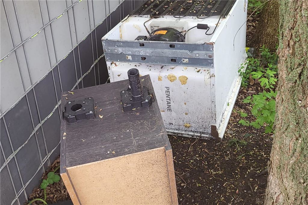 Große Elektrogeräte und Möbelstücke liegen hinter einem Zaun.