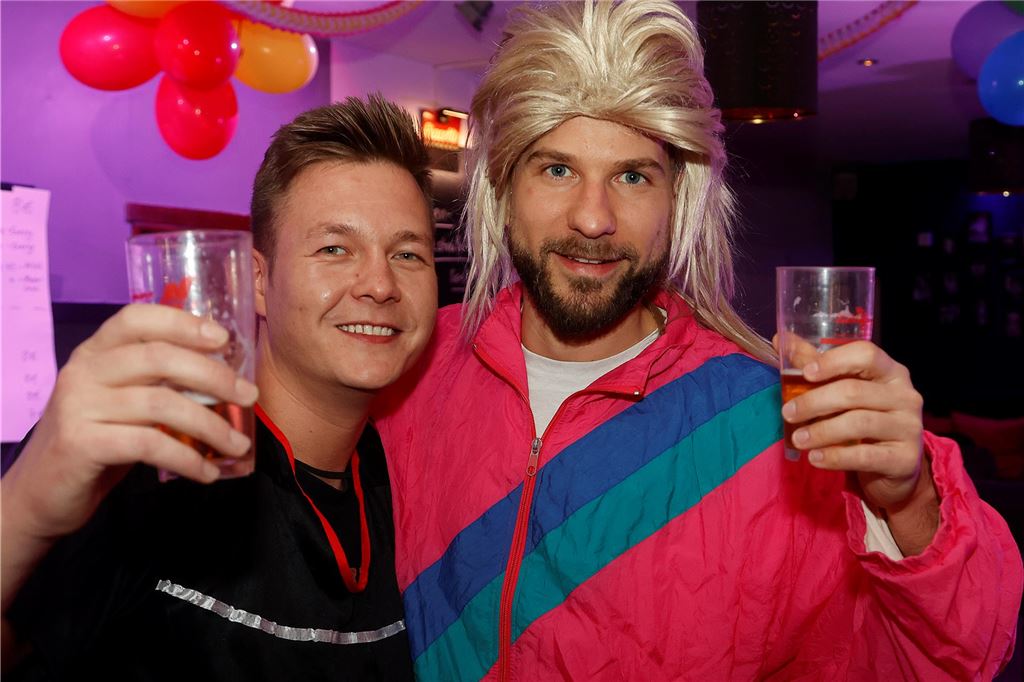 Zwei Männer halten ein Bier, der eine trägt einen pinken Jogginganzug und eine blonde Perücke.