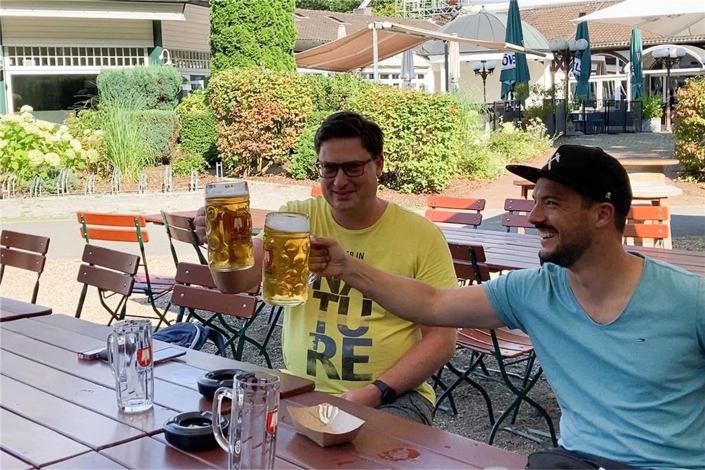Traditionelle Biergärten im Dortmunder Süden: Alle Infos im Überblick Zwei Männer sitzen an einem Tisch im Freien und stoßen mit Bierkrügen an. Die Szene spielt sich vor dem Hintergrund einer gepflegten Außenanlage ab.