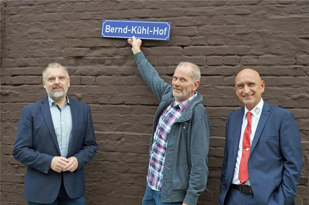 Neue Kita und OGS-Räume an der Schillerschule in Massen eröffnet Unnas Bürgermeister Dirk Wigang (von links nach rechts), der Hausmeister der Schillerschule und der Schulleiter Matthias Landsberg stehen vor einer braun gestrichenen Mauer. Der Hausmeister hält ein Schild mit der Aufschrift "Bernd-Kühl-Hof" hoch.