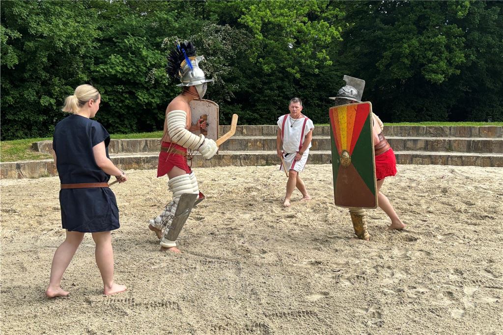 Zwei Gladiatoren kämpfen. Daneben steht der Gladiatorenmeister und eine Schiedsrichterin. 
