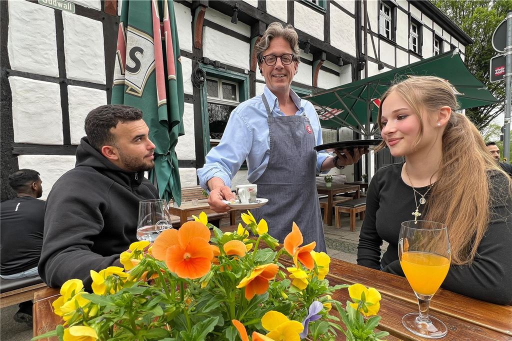 Unnas Biergärten starten in die Saison 2025 Vor der Gaststätte Meisterhaus in Unna serviert Wirt Daniel Gala einer Frau und einem Mann am Tisch einen Kaffee. Vor ihnen stehen Blumen.