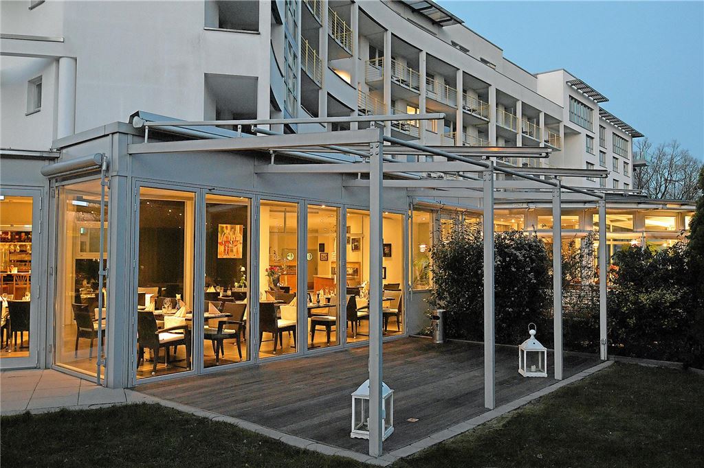 Buffet und Brunch zu Ostern: Diese Restaurants haben besondere Menüs Das Bild zeigt ein Restaurant mit Glasfront und einer überdachten Terrasse. Im Innenraum des Restaurants sind gedeckte Tische und Stühle zu sehen.