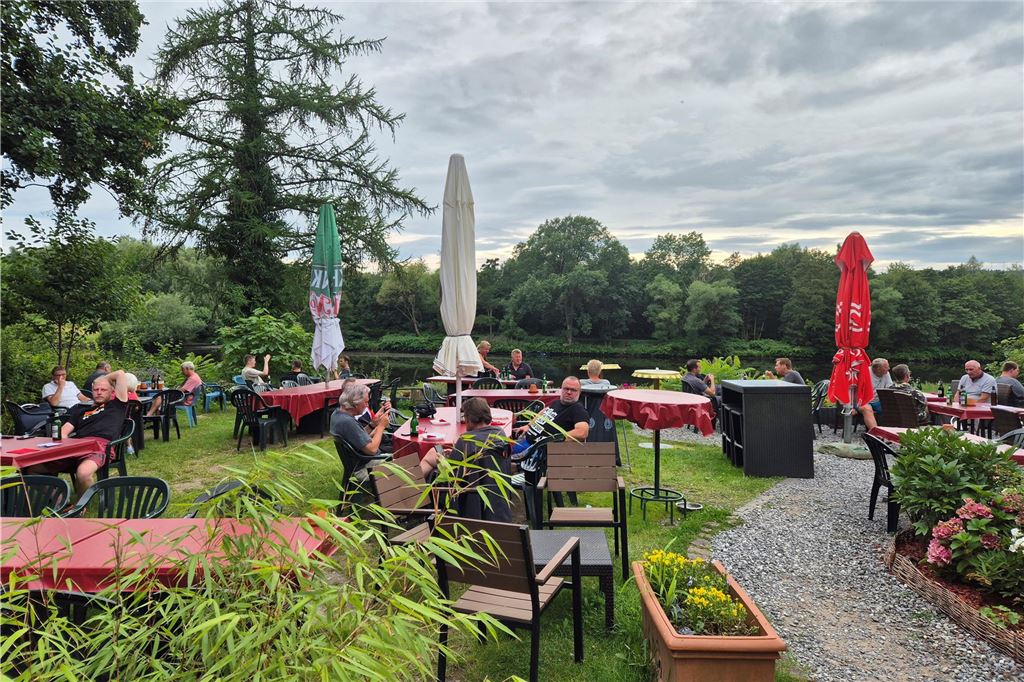 Traditionelle Biergärten im Dortmunder Süden: Alle Infos im Überblick Auf dem Bild sind im Freien sitzende Personen zu sehen, die sich an rot gedeckten Tischen eines Biergartens aufhalten. Im Hintergrund befindet sich ein Gewässer und Bäume, während der Himmel bewölkt ist.