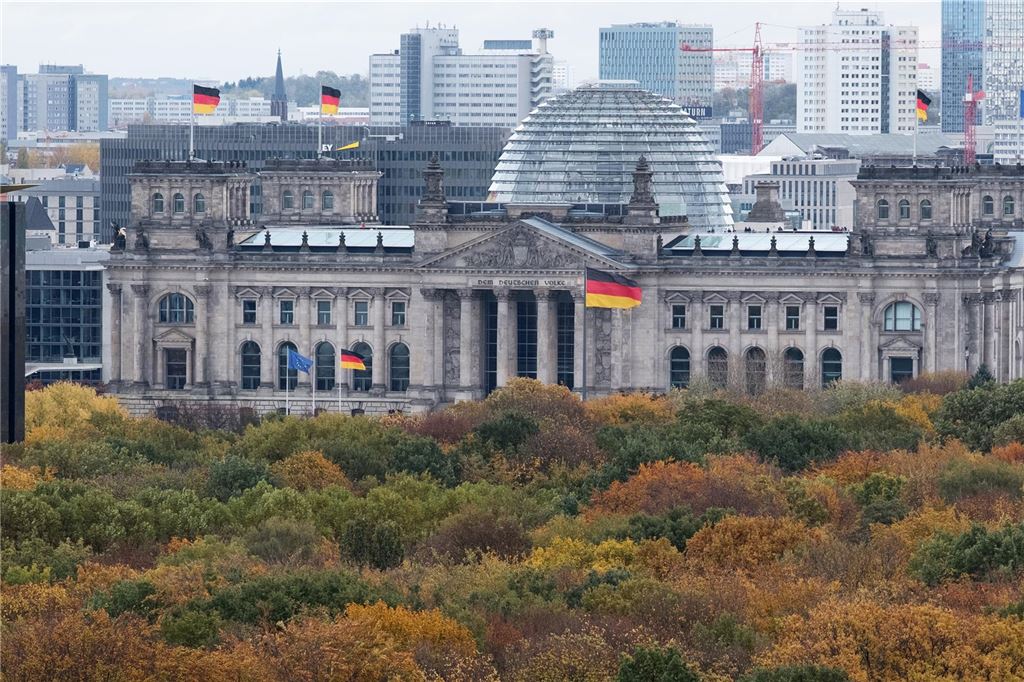 Unnas Bahnhof hat ICE-Anschluss – vorübergehend Der Reichstag in Berlin mit seiner bekannten Glaskuppel, fotografiert über das herbstlich eingefärbte Laub eines Waldstücks hinweg und mit Wolkenkratzern im Hintergrund.