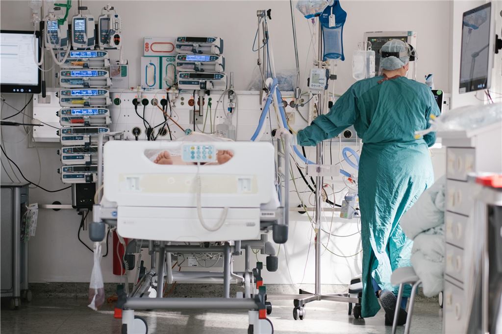 Staatsanwaltschaft ermittelt nach Tod einer Patientin (22) Das Symbolbild zeigt eine Situation auf einer Intensivstation. Ein Patient liegt mit nackten Füßen im Intensiv-Bett. Eine OP-Schwester steht daneben und schaut auf einen Bildschirm.