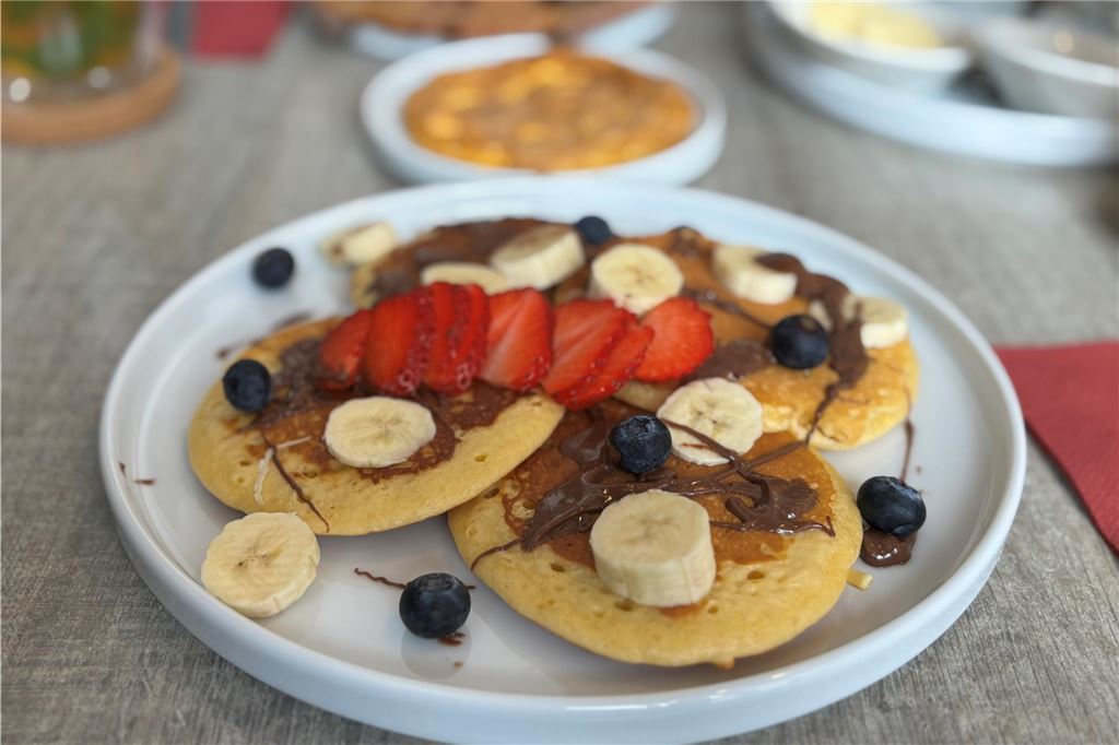 Restaurant-Check: Bei „Amlounge“ kann man den ganzen Tag frühstücken Auf dem Foto ist ein Teller mit Pancakes zu sehen, die mit Schokoladencreme, Bananen, Blaubeeren und Erdbeeren belegt sind. Im Hintergrund befinden sich weitere Teller mit Speisen und ein Glasgefäß.