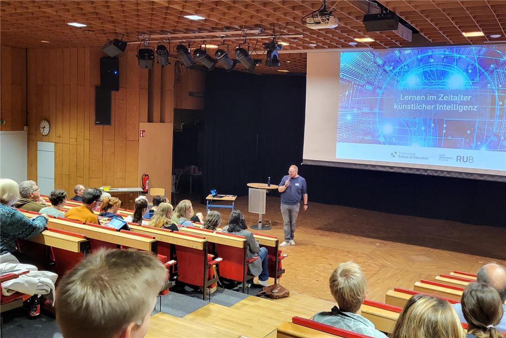 Schulung in der Gesamtschule: So verändert Künstliche Intelligenz den Unterricht In einem Hörsaal sitzen etwa zwei Dutzend Lehrerinnen und Lehrer im Publikum. Am Rednerpult steht Referent Matthias Kostrzewa. Hinter ihm ist eine Leinwand auf die seine Vortragspräsentation projiziert ist. Dort ist der Titel des Vortrags zu lesen: Lernen im Zeitalter künstlicher Intelligenz.