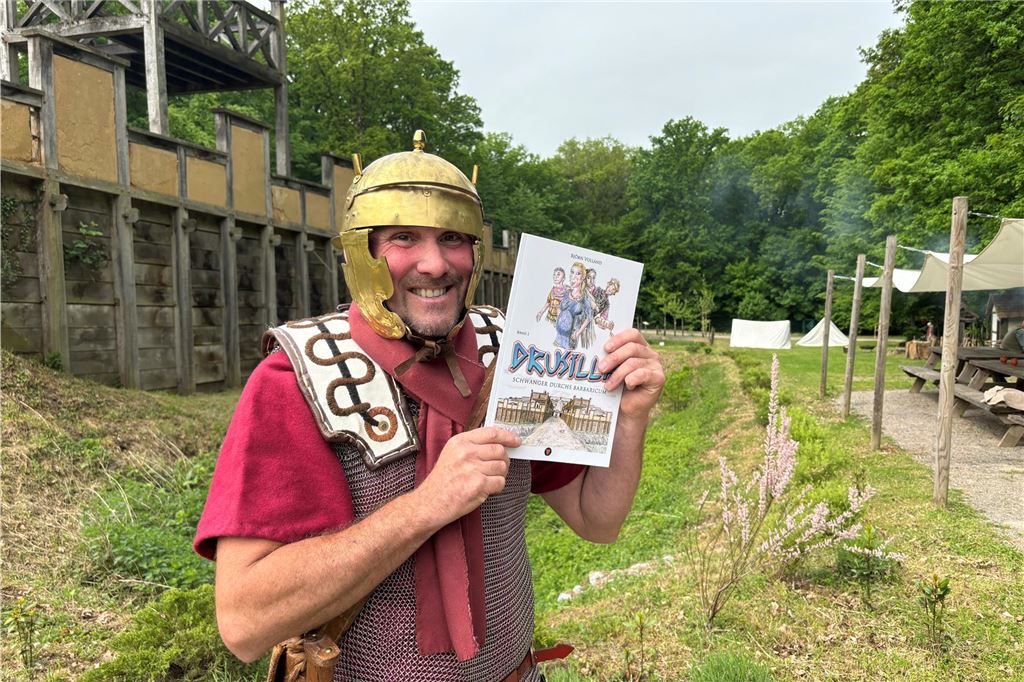 Björn Volland steht mit dem Comic "Drusilla - Schwanger im Barbaricum" vor der Holz-Erde-Mauer im Römerpark.