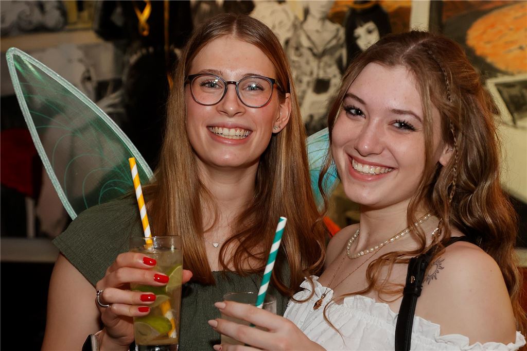 Zwei Frauen mit Feenflügeln und Cocktails lächeln in die Kamera.