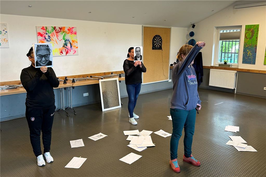 Arbeit und sich selbst finden: Theaterprojekt stärkt alleinerziehende Frauen Drei Teilnehmerinnen des Lebens:Art-Projekts stehen auf der Probenfläche. Zwei halten Masken mit Gesichtsausdrücken vor dem Gesicht. Eine erhebt ihren Arm, während sie etwas vorträgt.