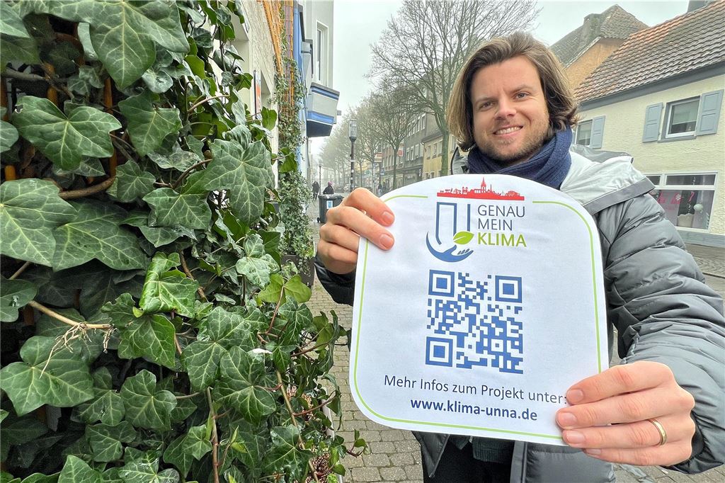 Ideen für Klimaschutz gesucht – erstmals Wirtschaftspreis „Grünes Unna“ Klimaschutzmanager Thomas Heer hält ein Hinweisschild zum Thema "Genau mein Klima" mit einem QR-Code.