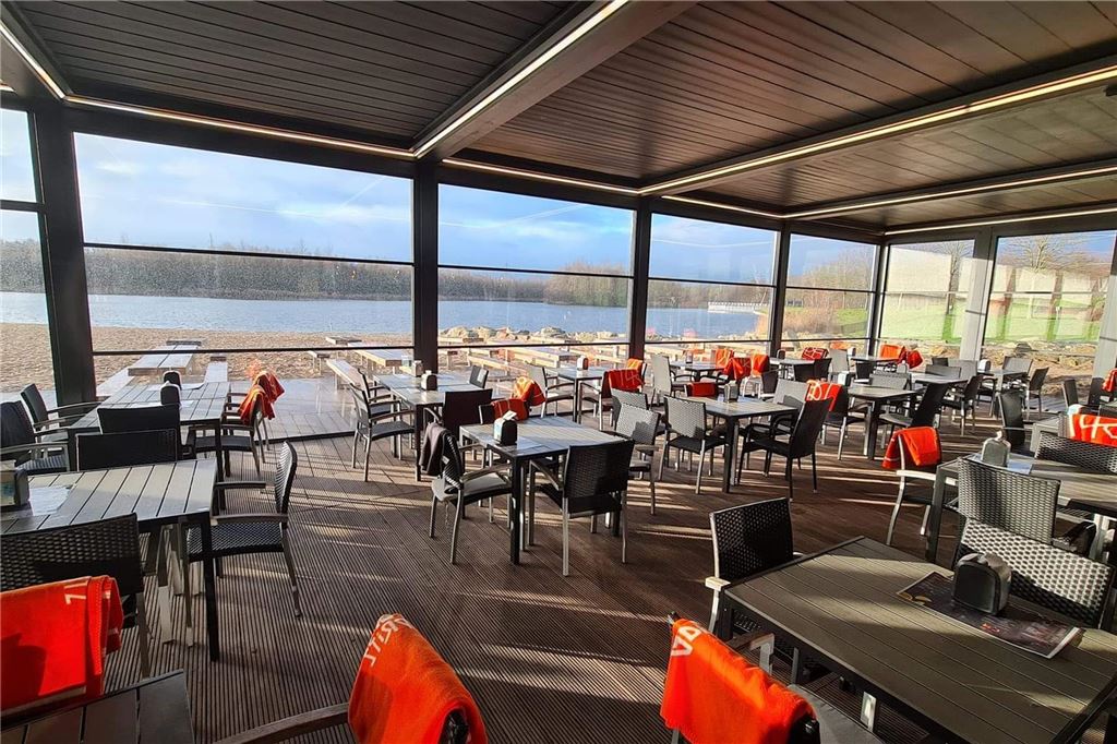 Buffet und Brunch zu Ostern: Diese Restaurants haben besondere Menüs Ein Innenbereich mit Tischen und Stühlen blickt durch Fenster auf einen See. Auf den Stühlen liegen Handtücher.