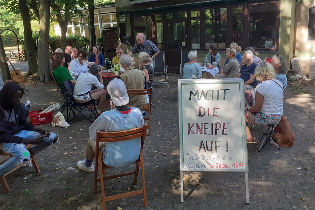 Planungsverfahren nötig vor Neueröffnung des Bistros Menschen sitzen vor der geschlossenen Gastronomie am Kurpark und fordern mit einem Schild "Macht die Kneipe auf".