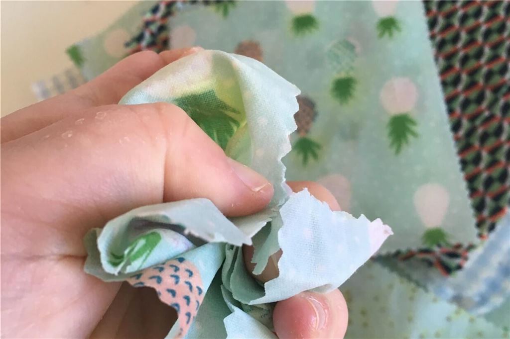 So macht man Bienenwachstücher selbst: Einfache Anleitung zum Nachbasteln Eine Hand knüllt das bunte Stück Stoff mit dem Wachs.