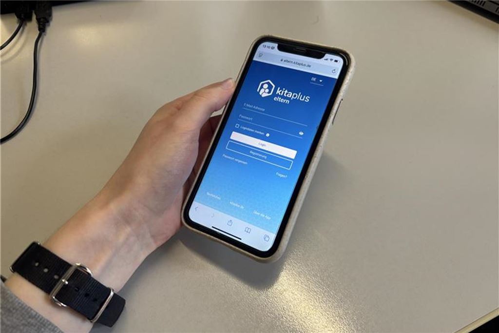 Schließtage und Notgruppen: Wie oft bleiben Kitas geschlossen? Ein Smartphone zeigt die Anmeldeseite der "kitaplus eltern" App. Die Hand einer Person hält das Telefon, während auf dem Display die Felder für E-Mail-Adresse und Passwort sichtbar sind.