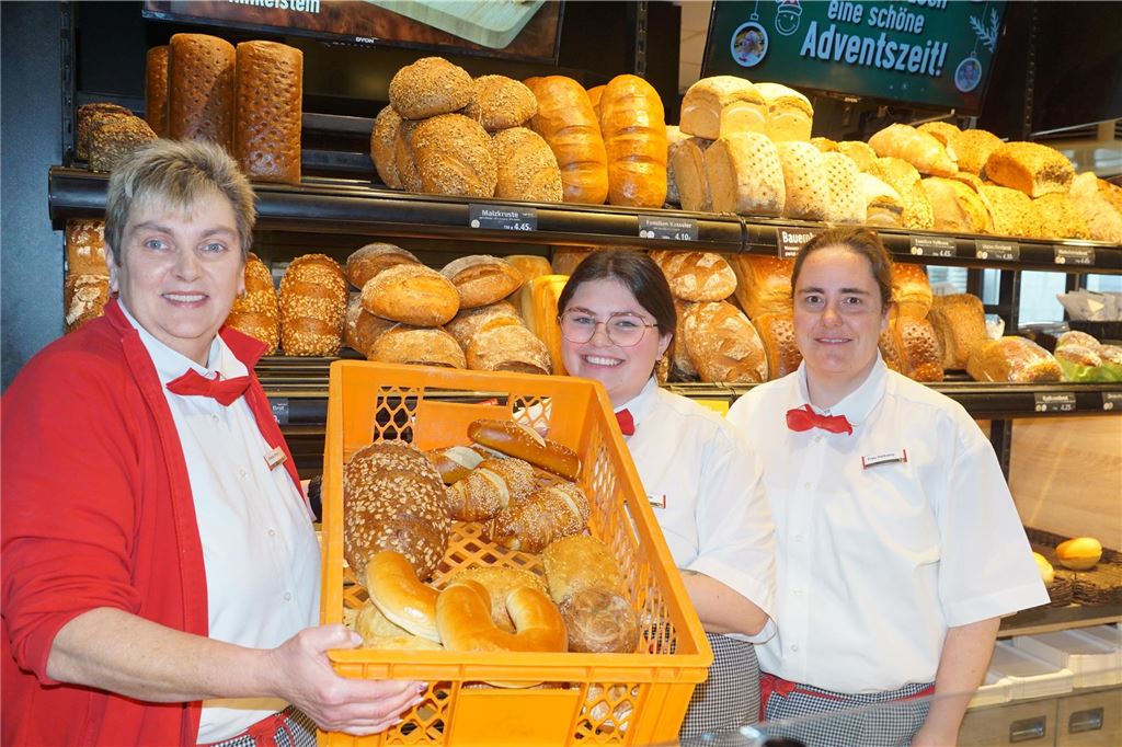 Hier gibt’s an den Feiertagen frische Brötchen Drei Bäckereifachverkäuferinnen stehen hinter der Brötchentheke und lächeln. Eine zeigt einen Korb mit verschiedenen Backwaren wie Brötchen und Milchhörnchen.