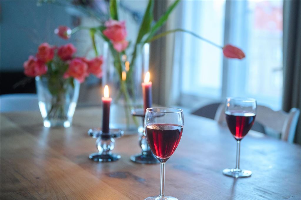 Restaurant-Aktionen zum Valentinstag: Romantische Menüs und Live-Musik Zwei Gläser Rotwein stehen auf einem Tisch in einem Wohnzimmer. Zwei Kerzen und zwei Vasen mit Blumen stehen ebenfalls auf dem Tisch.