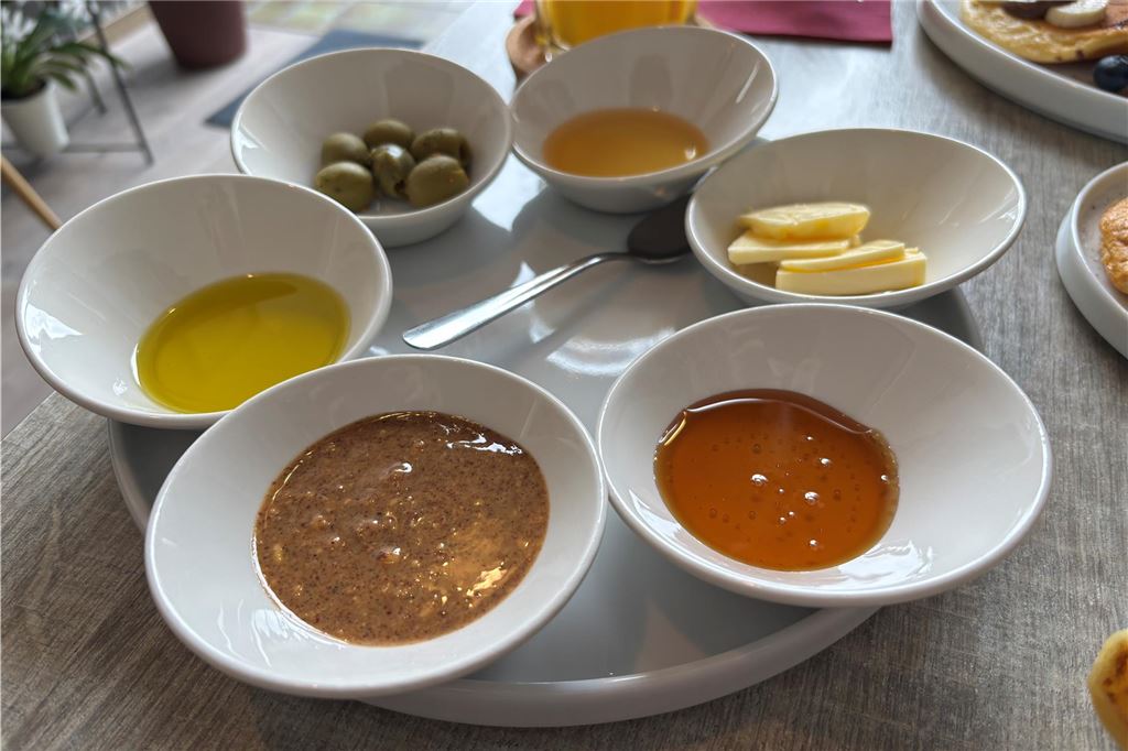 Restaurant-Check: Bei „Amlounge“ kann man den ganzen Tag frühstücken Das Foto zeigt eine Auswahl von Speisen, die in kleinen weißen Schalen auf einem Teller angerichtet sind. Zu sehen sind unter anderem Oliven, Honig, Butter, Amlou und Olivenöl, vermutlich als Teil eines Frühstücks.